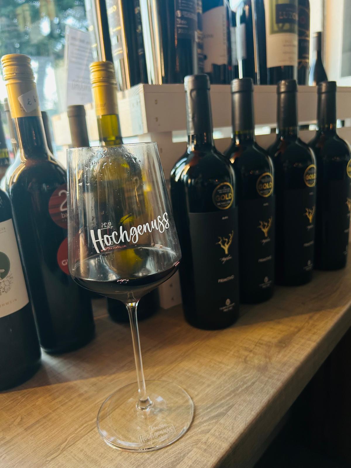 Ein Weinglas mit der Aufschrift "Hochgenuss". Im Hintergrund sind mehrere Flaschen Rotwein zu sehen.